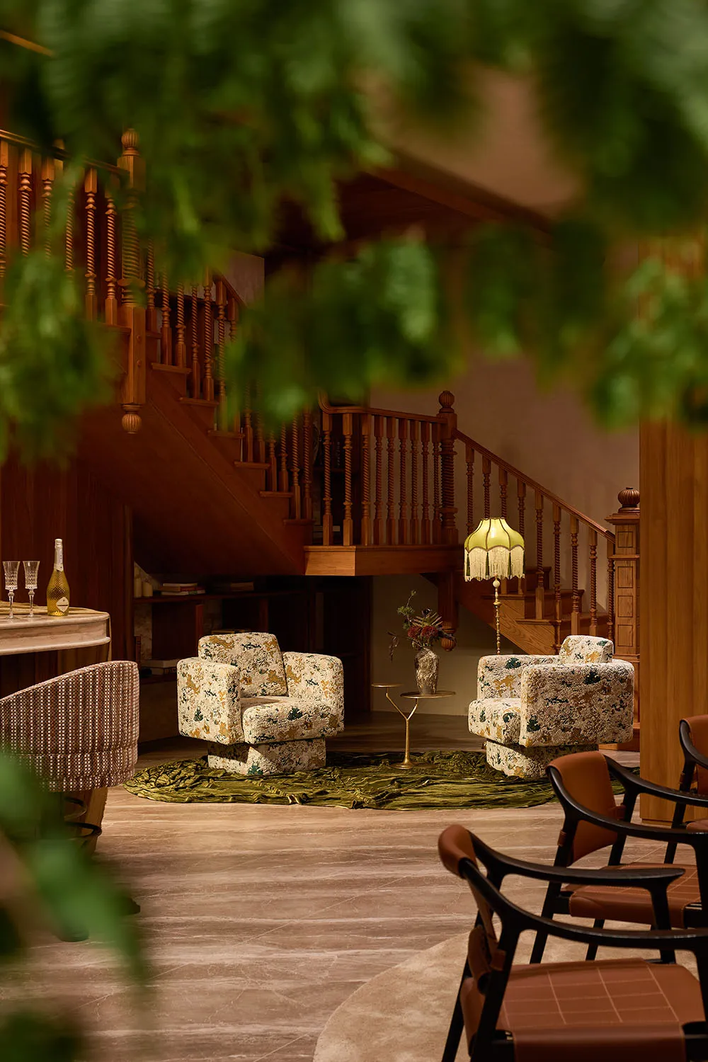 Interior view of a luxurious lounge area featuring two floral-patterned armchairs, a green velvet rug, wooden staircase with turned balusters, and warm ambient lighting from a fringed floor lamp.