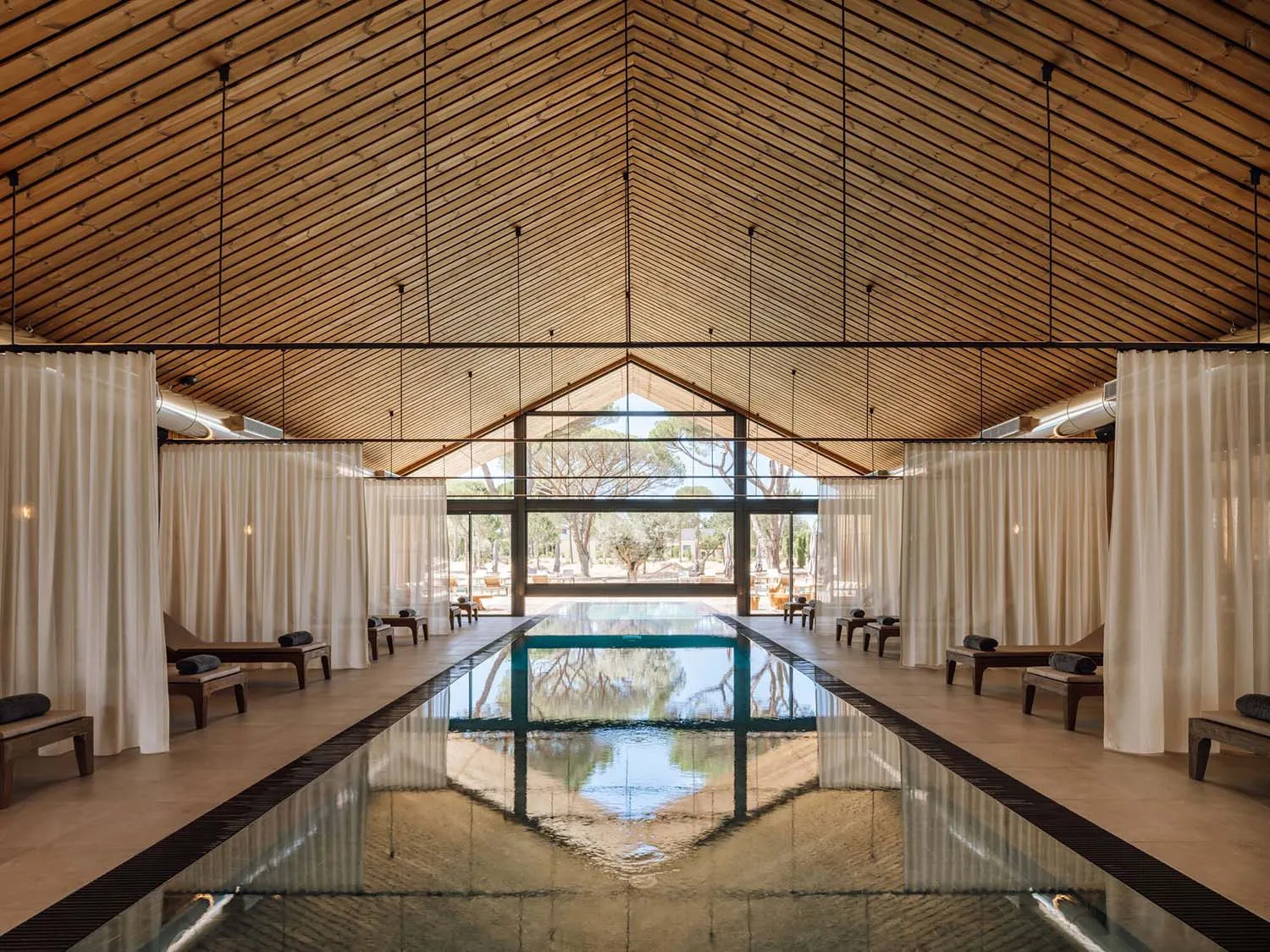 Long indoor lap pool in a serene spa with high vaulted wooden ceiling, white curtains, and wooden lounge chairs on both sides.