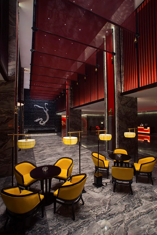 Luxurious hotel lobby featuring grey marble floors, yellow velvet armchairs, black tables, dramatic red ceiling panels, and a large white dragon mural on a black stone wall.
