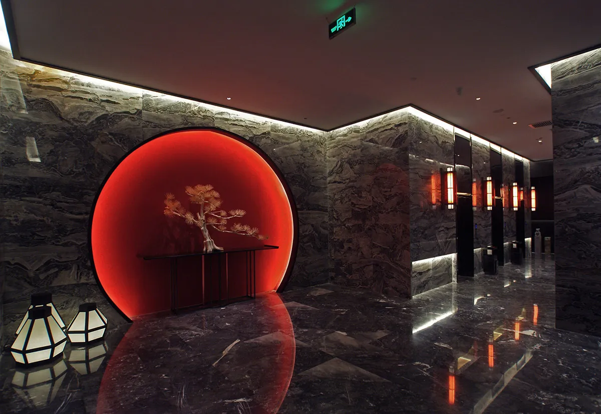 Modern luxury hotel lobby with dark marble walls, a large illuminated red circular feature wall containing a console table with bonsai tree, and geometric lanterns on the glossy floor.