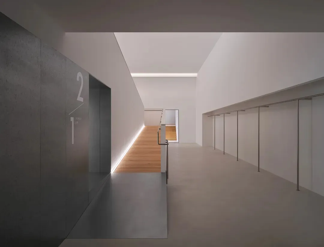 Modern minimalist interior hallway with grey concrete walls, light oak flooring, recessed LED lighting, and a view of a floating staircase in the distance.