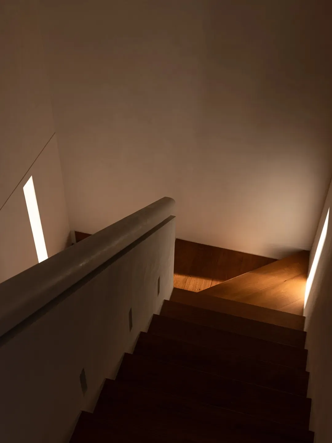 View looking down a modern wooden staircase with dark oak steps, smooth concrete-like walls, and warm LED strip lighting integrated along the sides.