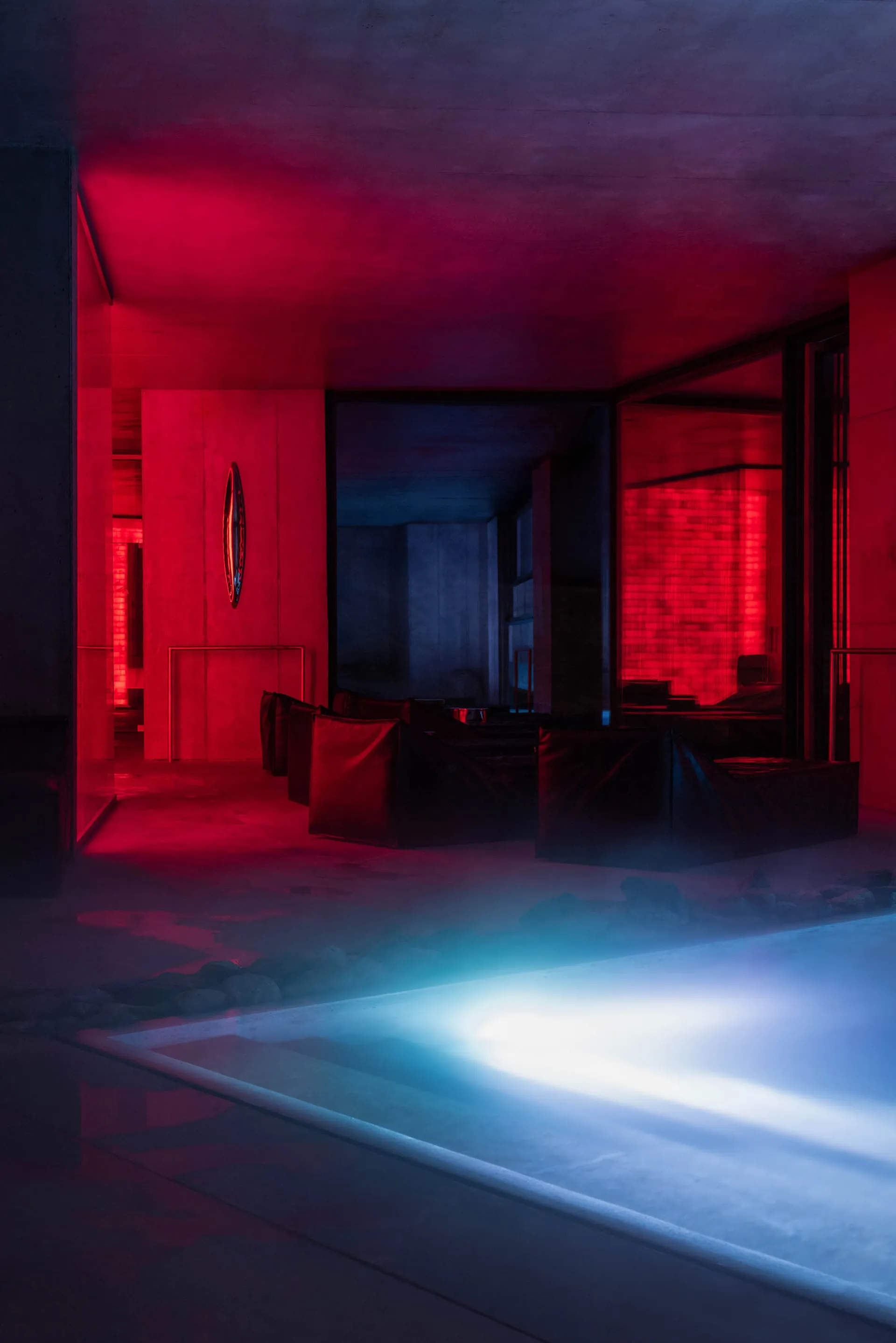 Modern indoor swimming pool surrounded by concrete architecture, featuring modular dark lounge seating, dramatic red uplighting on the ceiling and walls, and bright blue underwater lighting in the pool.