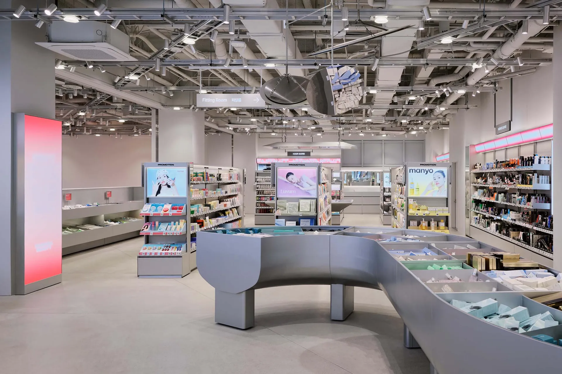 Interior view of a modern cosmetics and skincare store featuring a large curved grey display island, product shelving, digital advertisements, and exposed ceiling infrastructure.