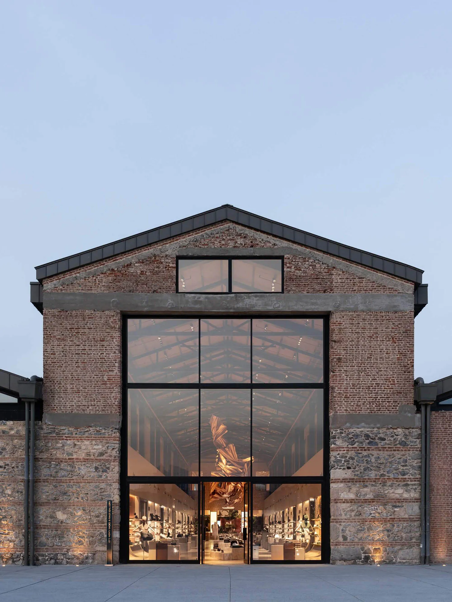 Front exterior view of a luxury retail store with a large black-framed glass facade set within a restored red brick and stone industrial building at dusk.