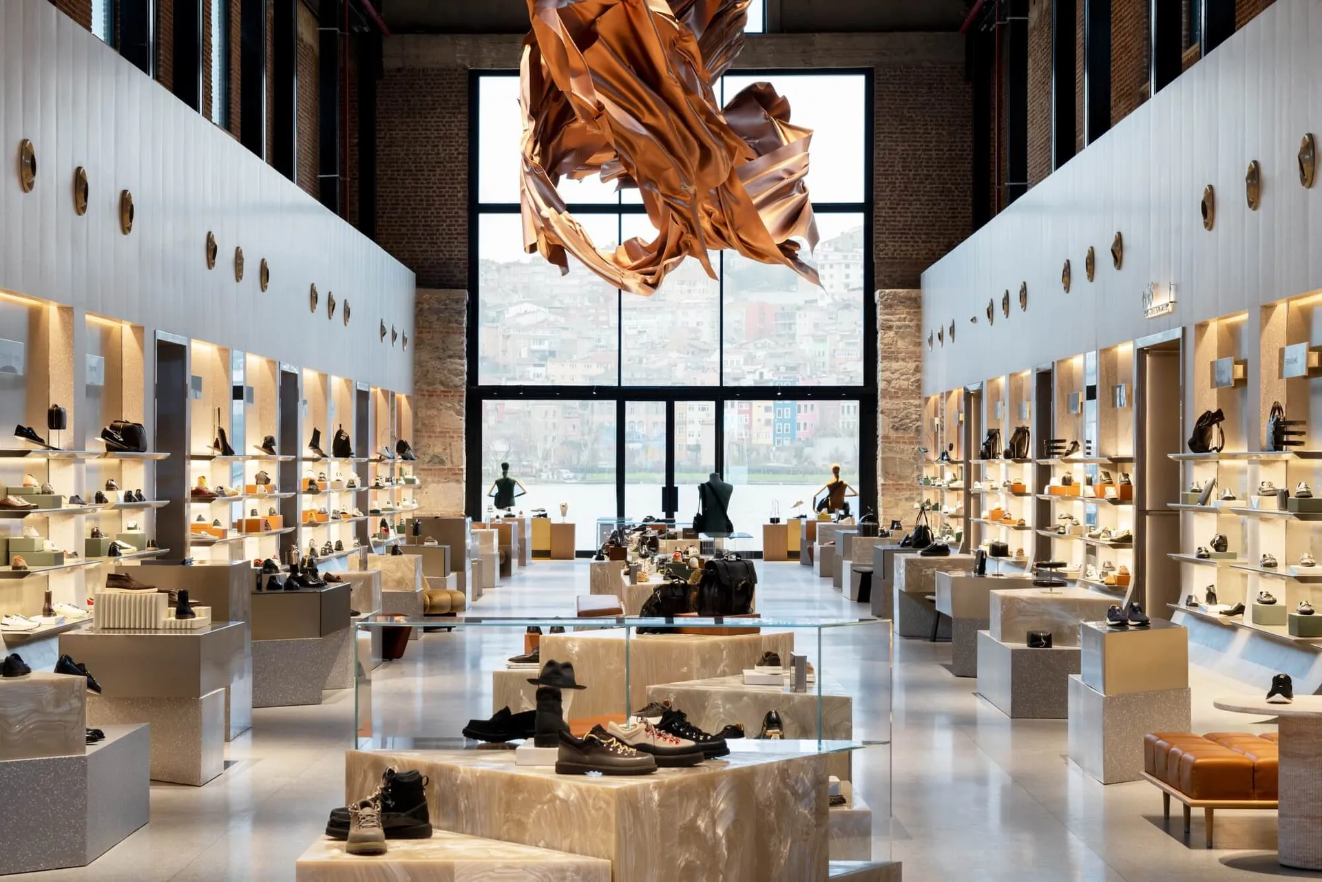 Wide view of a modern luxury shoe and handbag store featuring white walls, exposed brick, terrazzo display platforms, and a large abstract copper sculpture hanging from the ceiling with a cityscape visible through tall windows.