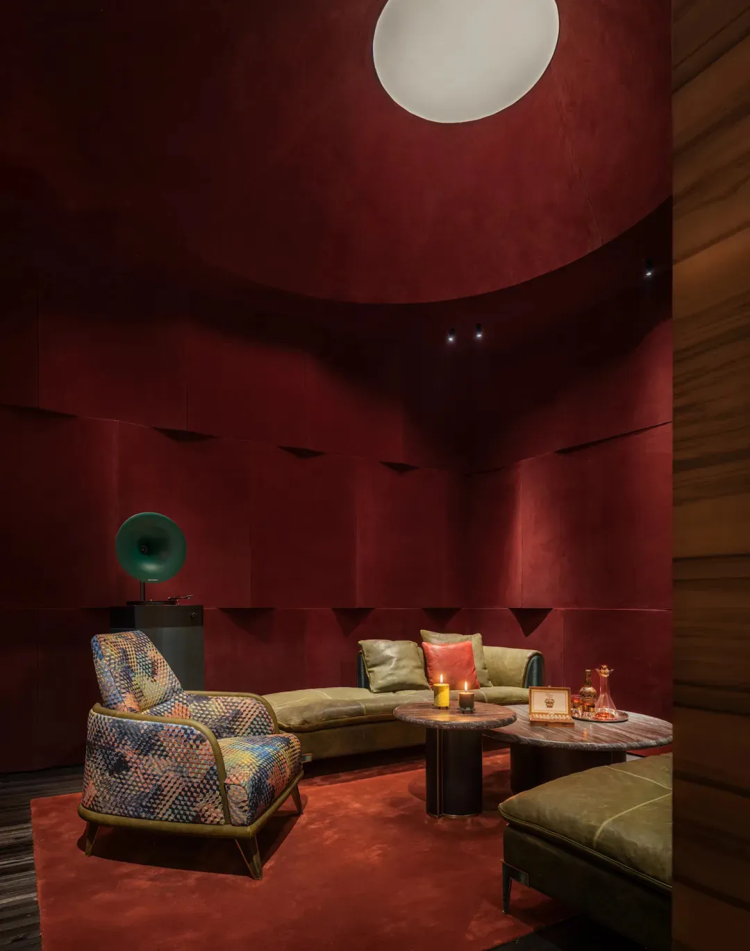 Modern lounge with curved red textured walls, patterned armchair, green leather sofa, round marble coffee tables, and large spherical pendant light.