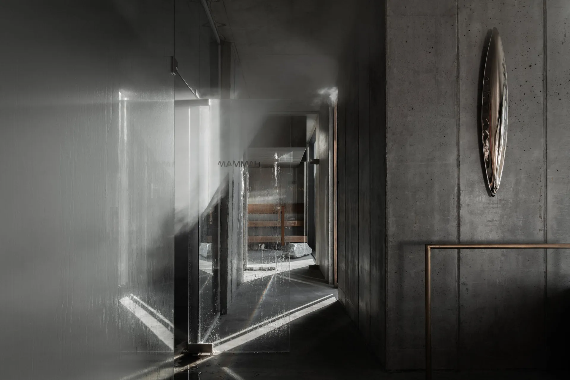 Narrow brutalist concrete hallway featuring a large translucent glass partition wall, dramatic diagonal light beams, and an asymmetrical metallic wall sculpture.