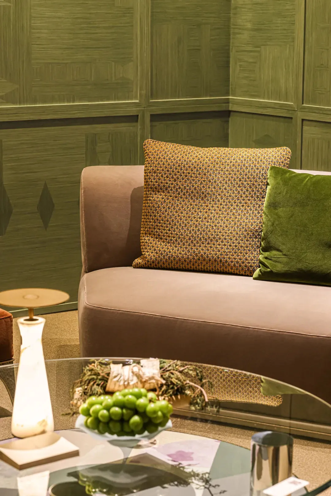 Close-up of a modern living room corner with a taupe velvet sofa, mustard patterned pillow, green velvet pillow, and an olive green textured wall panel background. A glass coffee table in the foreground holds a bowl of green grapes and a marble lamp.