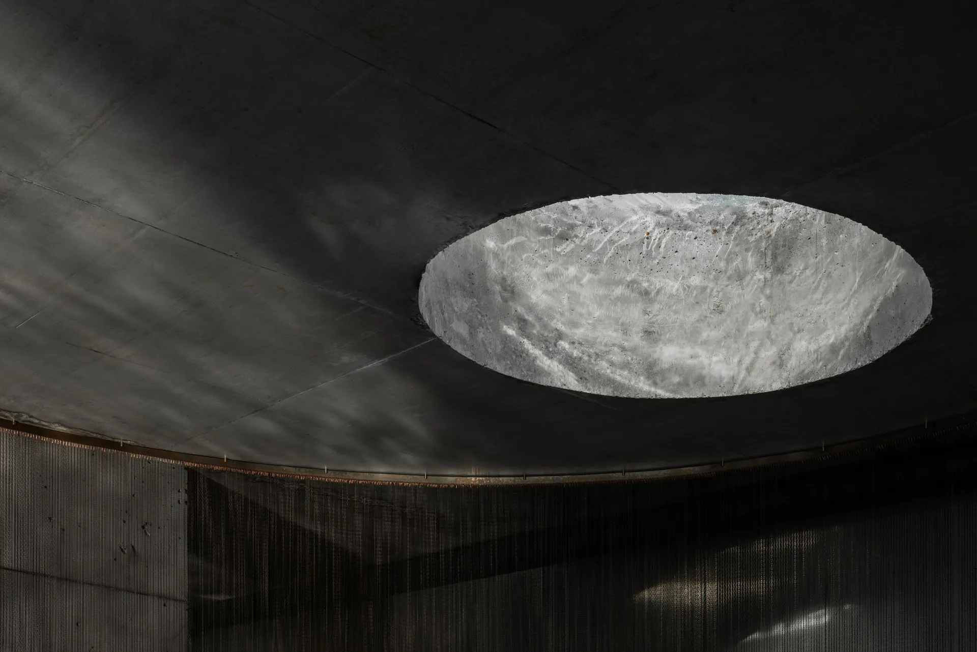 View looking up at a large circular concrete oculus skylight set into a dark raw concrete ceiling with hanging chainmail curtain below.