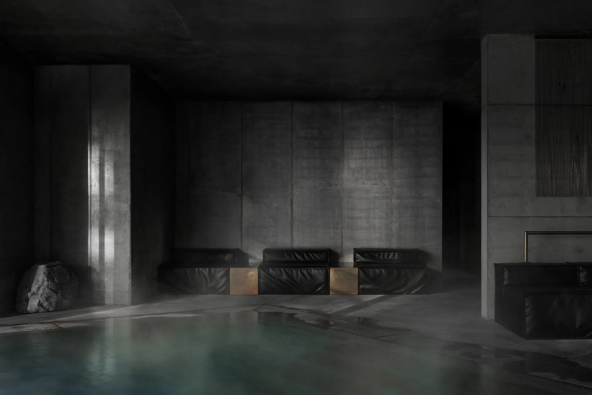 A dimly lit brutalist indoor swimming pool surrounded by massive concrete walls, featuring black leather modular benches with bronze accents and a large natural boulder on the left.