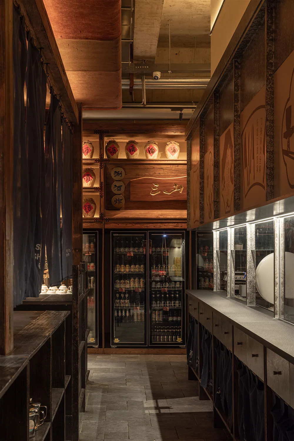 Narrow interior corridor of a Japanese izakaya featuring reclaimed wood shelves, hanging indigo noren curtains, illuminated ceramic sake jars, and glass-door refrigerators stocked with beverages.