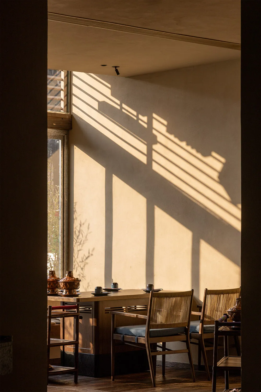 A tranquil wabi-sabi style dining room featuring wooden table and chairs with woven rattan backs, illuminated by warm sunlight that creates strong linear shadows on a textured plaster wall.