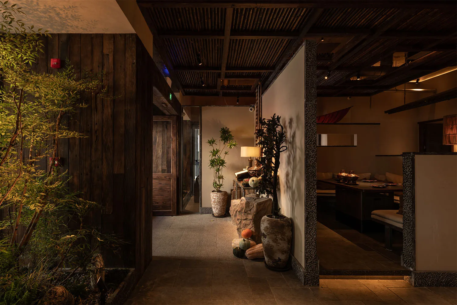 日式餐廳昏暗的走廊,深色木牆、質感石柱、盆栽植物,以及可看到溫暖用餐區與長椅座位的視野。