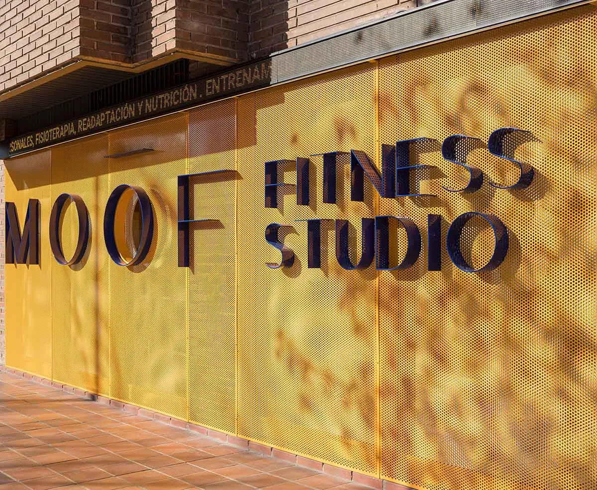 Exterior view of MOOF Fitness Studio featuring large 3D blue lettering mounted on a bright yellow perforated metal wall with terracotta tile flooring.
