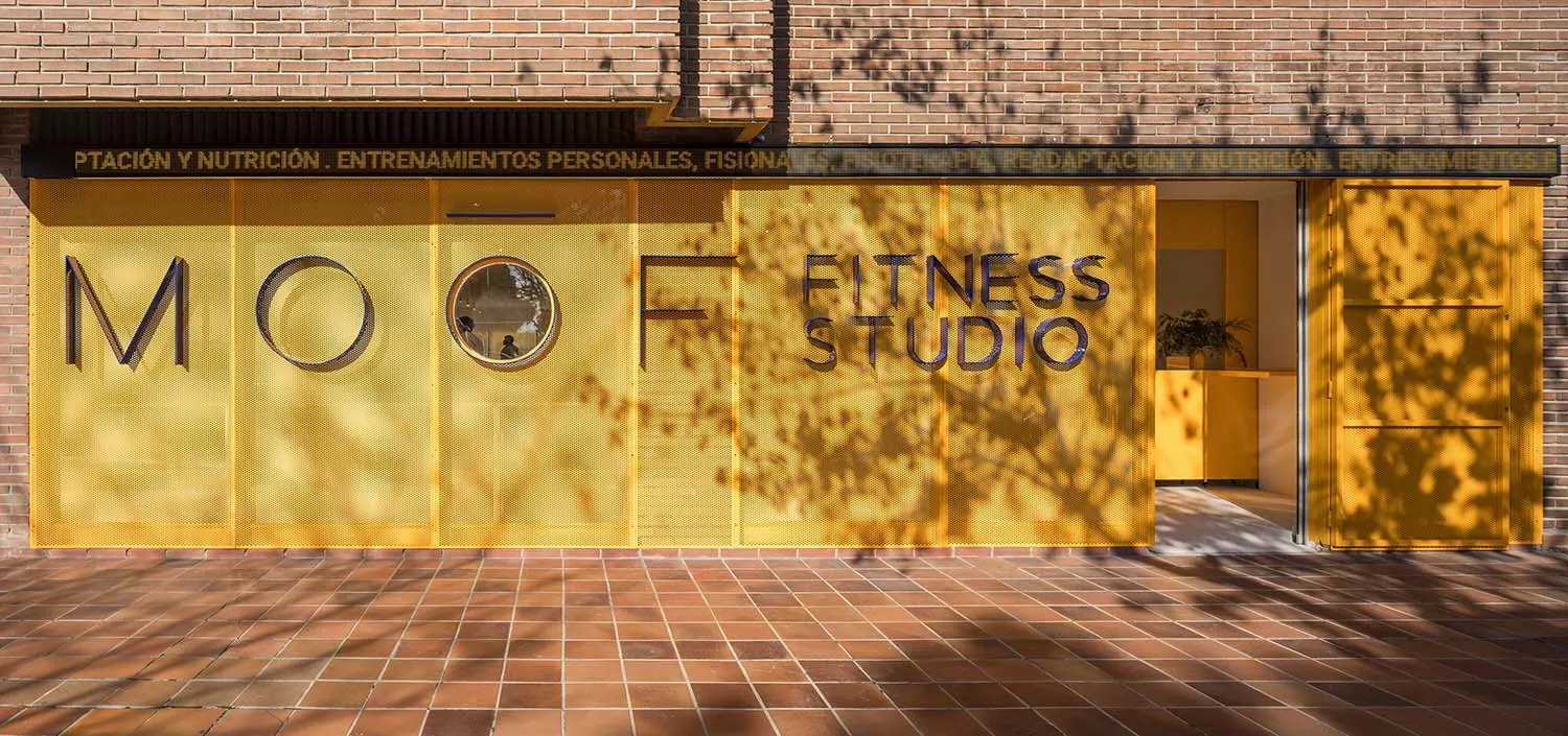 Modern fitness studio entrance with bright yellow perforated metal facade, large signage reading MOOF FITNESS STUDIO, and terracotta tile pavement in front of a brick building.