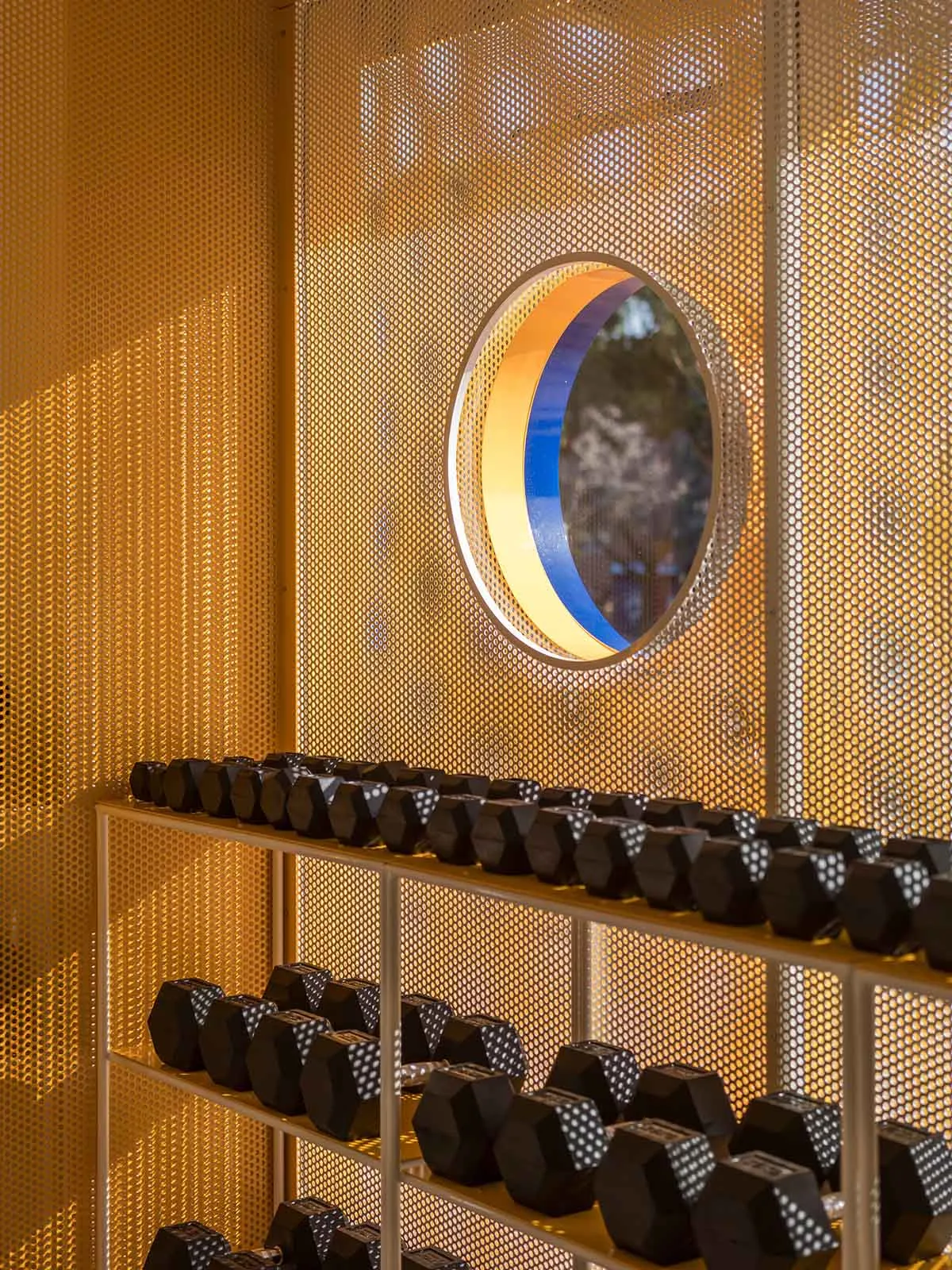 Modern home gym interior featuring a white metal dumbbell rack filled with black hexagonal dumbbells, set against a glowing golden perforated metal screen with a circular multicolored window.