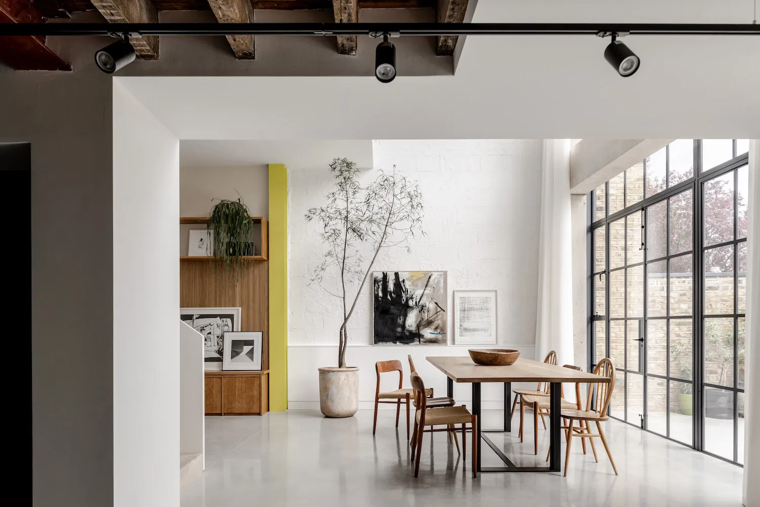 現代工業風格 loft 餐廳,長型橡木餐桌搭配多款木製餐椅、大型盆栽樹木、白磚牆,以及引入充足自然光的大型黑色格窗。