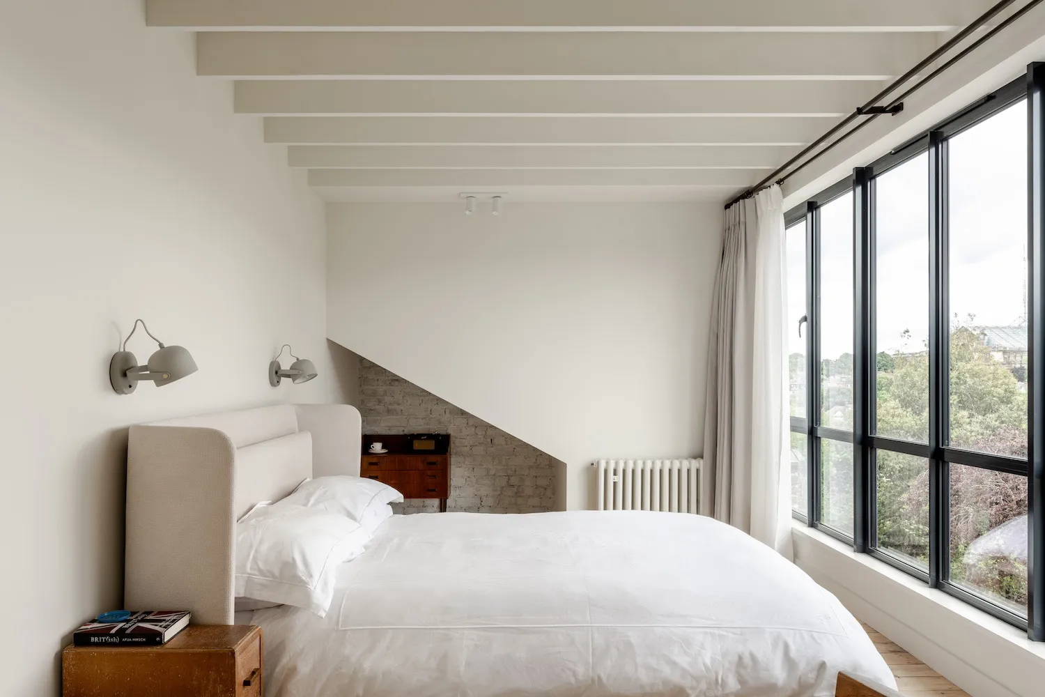 Minimalist bedroom interior with white walls, exposed brick under a sloped ceiling, large black-framed windows with sheer curtains, upholstered bed with white linens, wooden nightstand, and wall-mounted reading lamps.