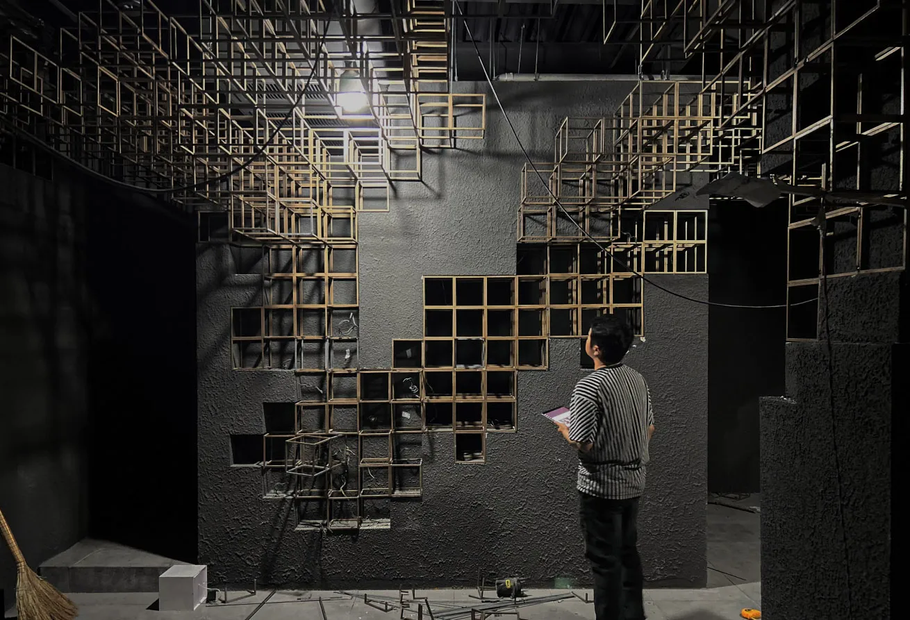 Man in striped shirt observing a large-scale wooden cube grid installation mounted on a dark concrete wall in an industrial exhibition space with dramatic overhead lighting.