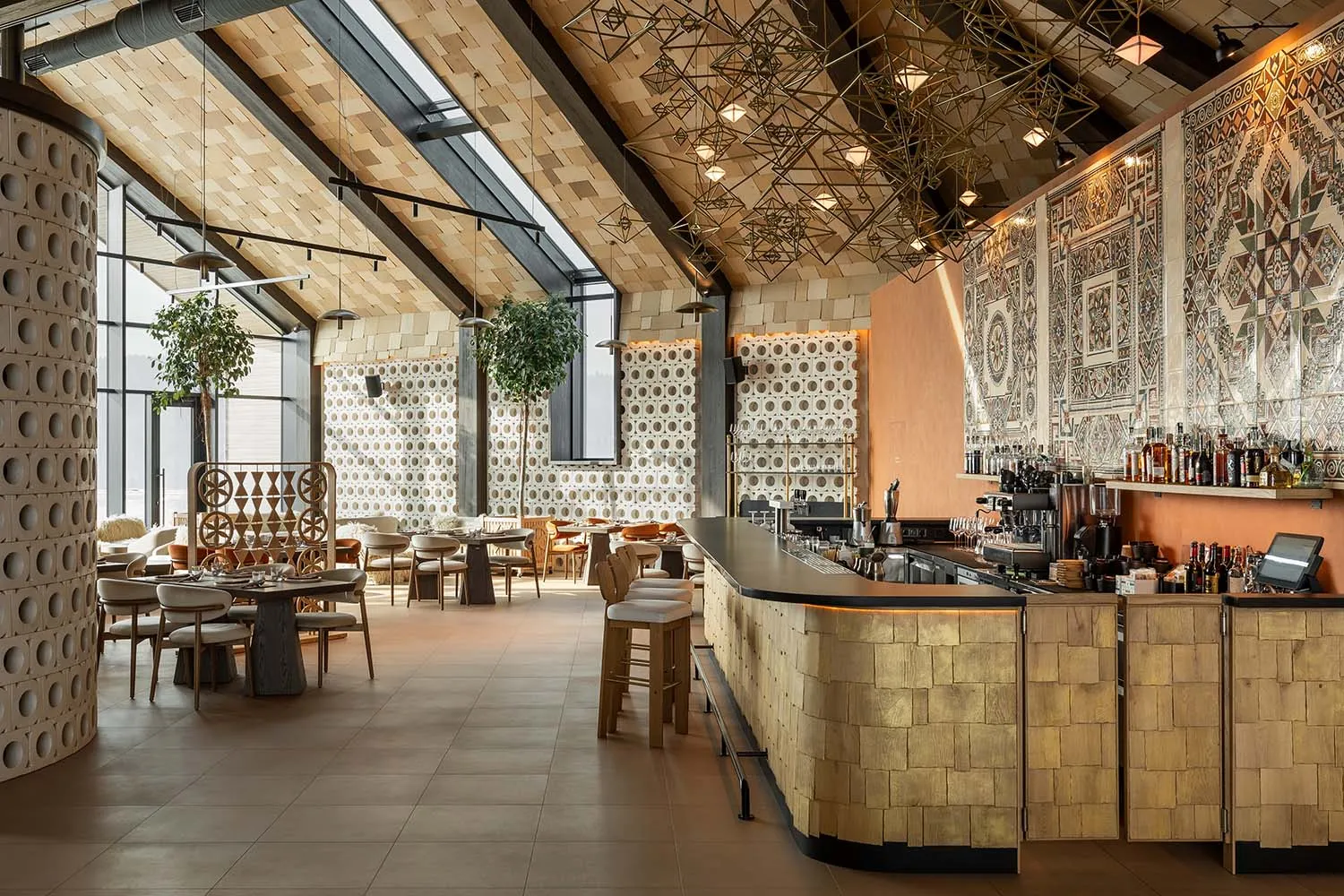 Modern restaurant interior with a curved terracotta-tiled bar, geometric brass pendant lights, patterned tile walls, wooden ceiling, and dining tables with neutral chairs.