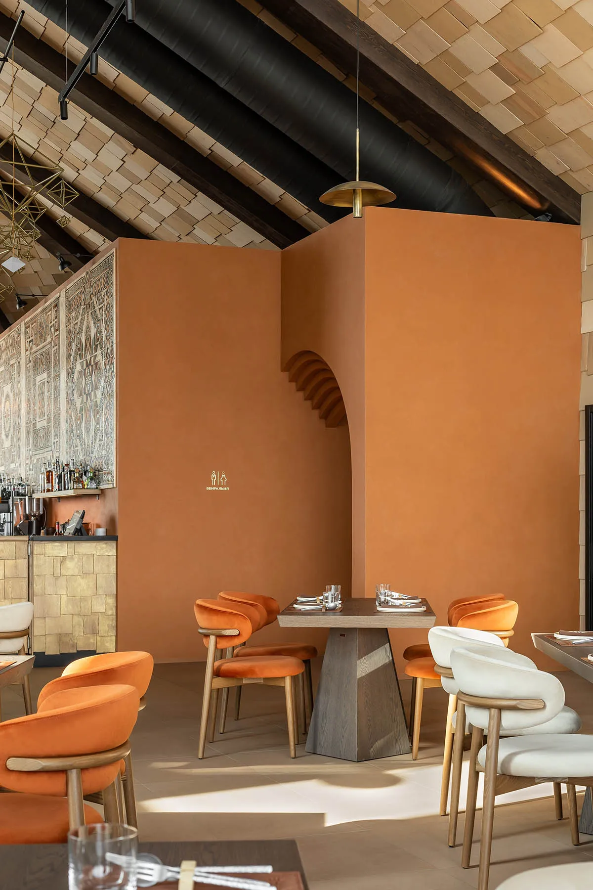 Modern restaurant interior with terracotta plaster walls, burnt orange and cream velvet dining chairs, wooden tables, and a striking high ceiling with exposed dark beams and wooden slats.