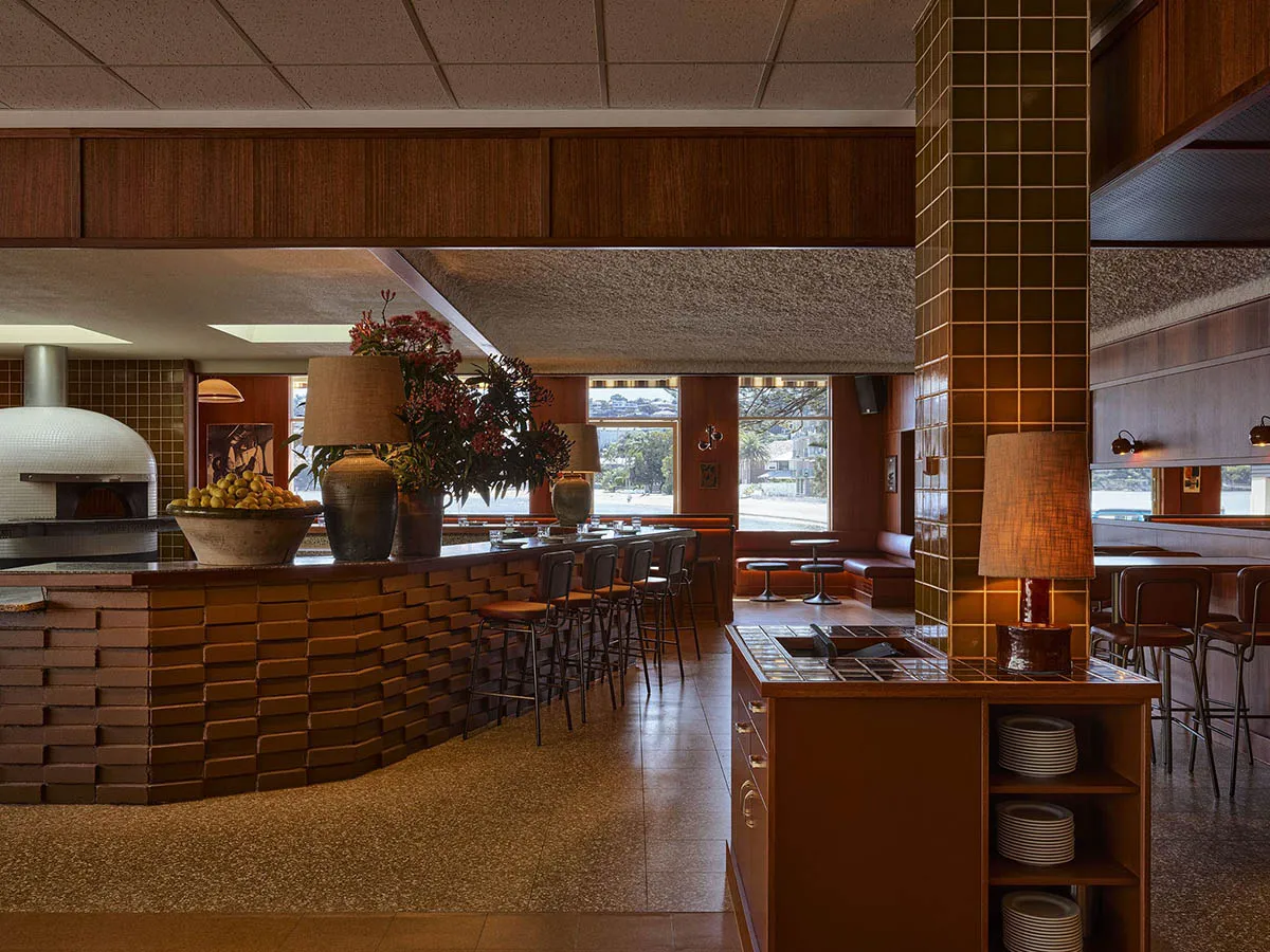 Interior view of a retro-style restaurant with a curved terracotta brick bar, wood-fired pizza oven, warm wood paneling, and leather bar stools under soft lighting.