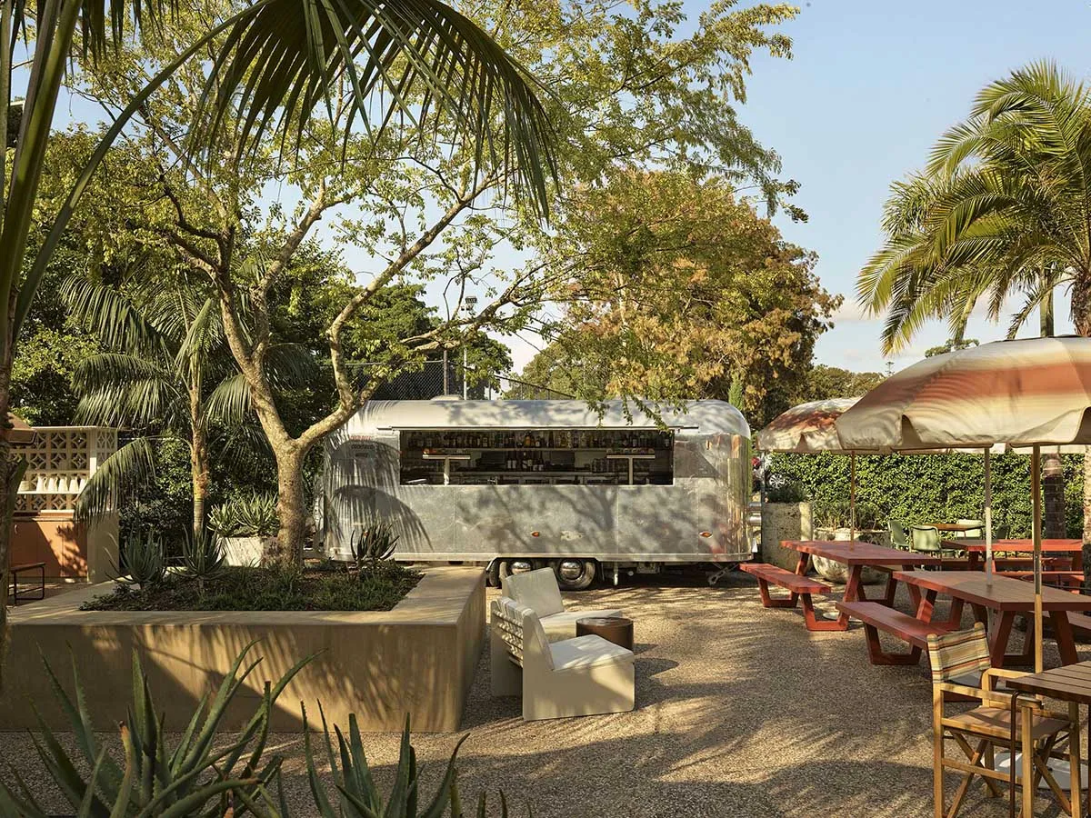 Outdoor tropical garden lounge with a vintage silver Airstream trailer converted into a bar, surrounded by palm trees, concrete planters with aloe and succulents, wooden picnic tables, and modern outdoor seating under a large striped umbrella.