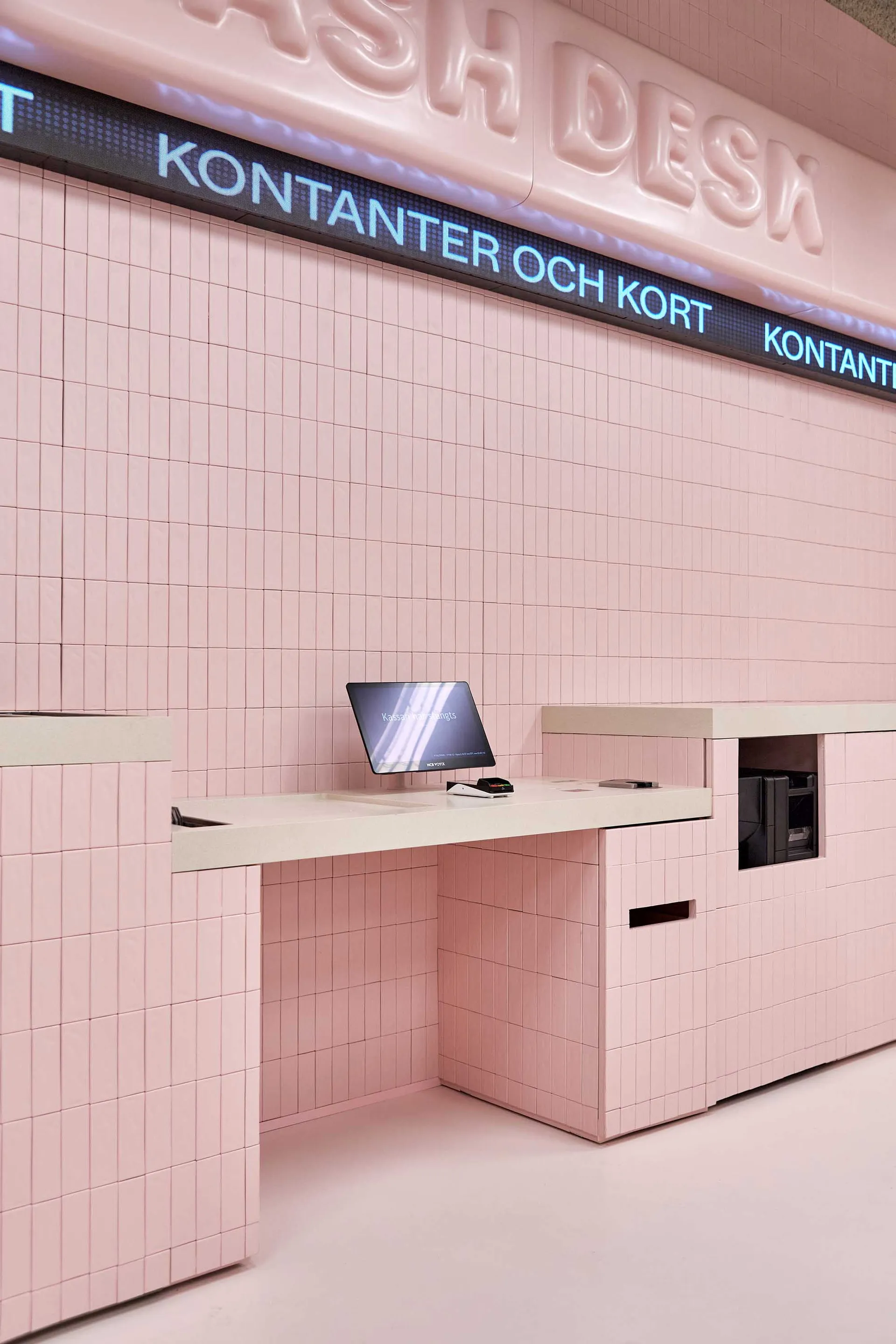 Modern cash desk covered in glossy pink square tiles with a built-in monitor and digital signage reading 'KONTANTER OCH KORT' against a matching pink tiled wall.