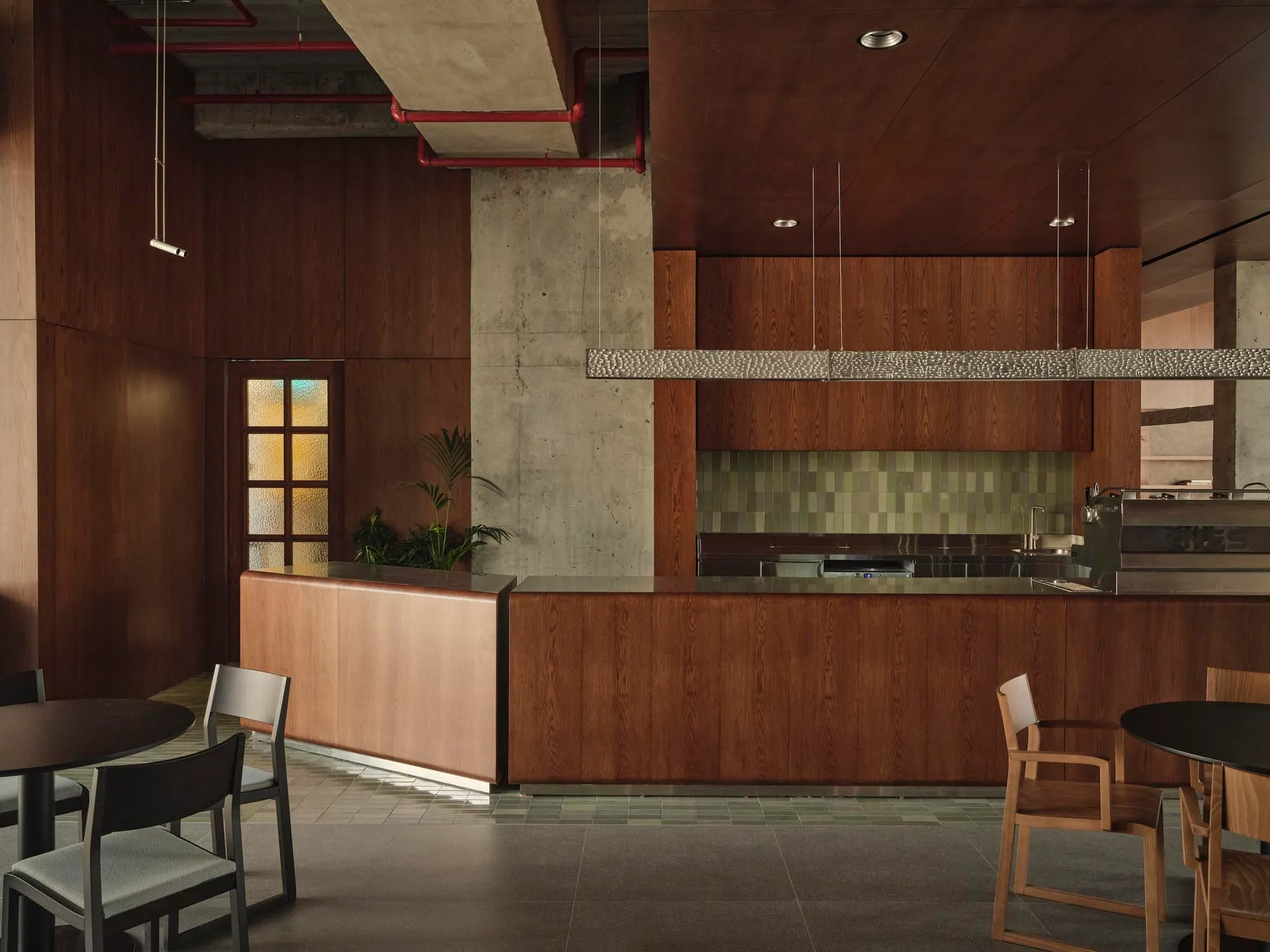 Modern industrial loft kitchen featuring a large walnut wood island, green tiled backsplash, concrete walls and ceiling with exposed red pipes, and wooden dining chairs under a hammered metal pendant light.