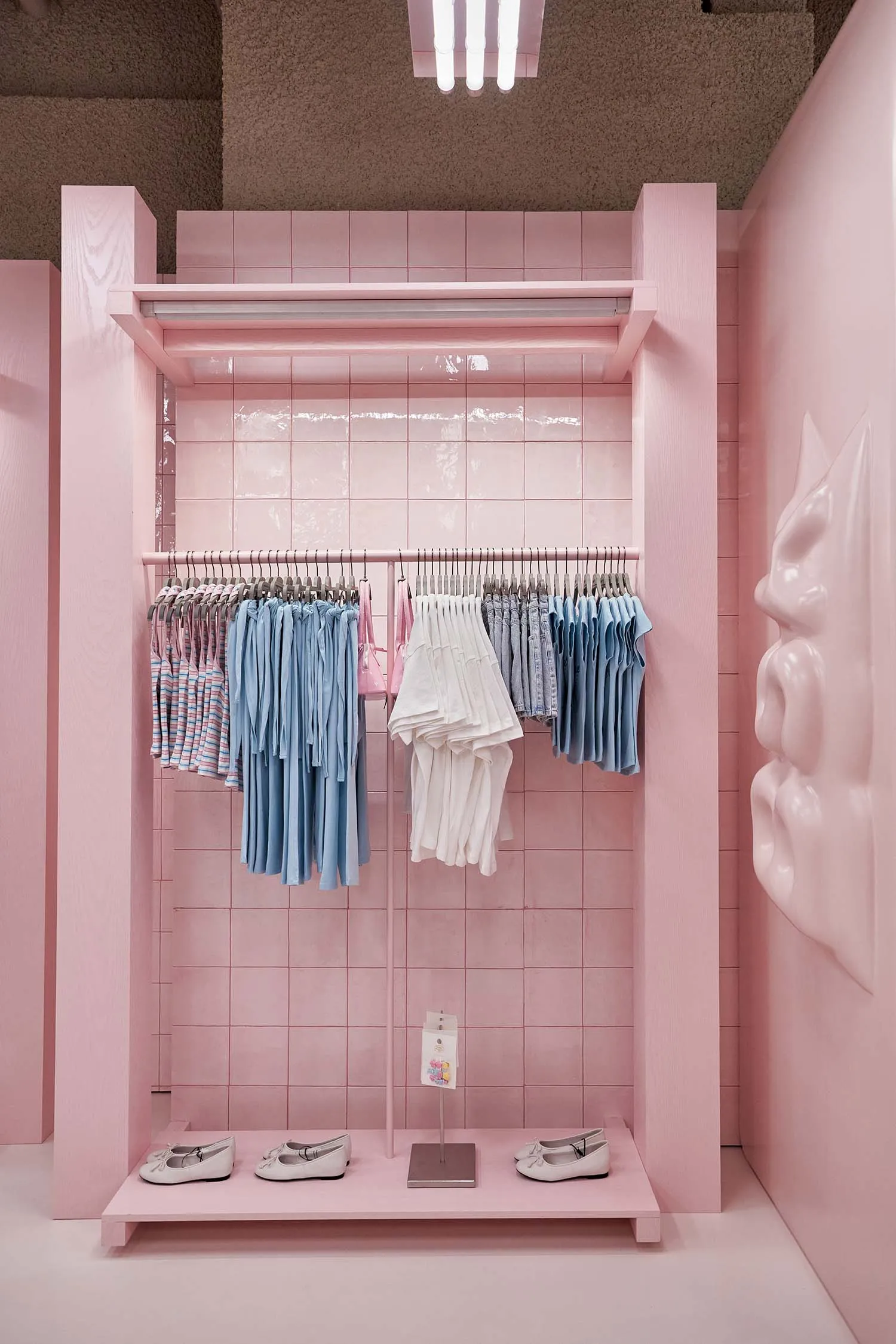 Pink tiled retail display alcove with clothing racks holding pastel and striped garments, white ballet flats on a pink platform, and a large sculptural pink face on the right wall.