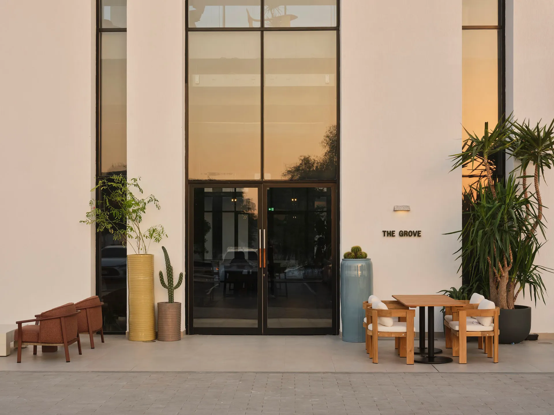 Modern minimalist building entrance with wooden armchairs, a square dining set, large colorful planters, and tall glass doors at dusk.