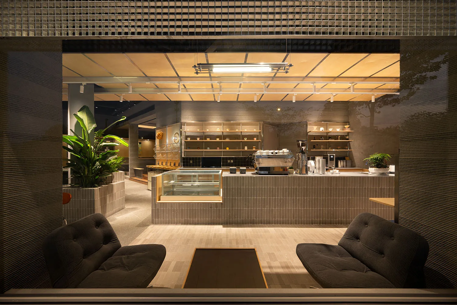 Modern minimalist cafe interior featuring two dark corduroy armchairs in the foreground, a low wooden table, ribbed concrete walls, a glass display case, and a professional espresso bar in the background under warm linear lighting.