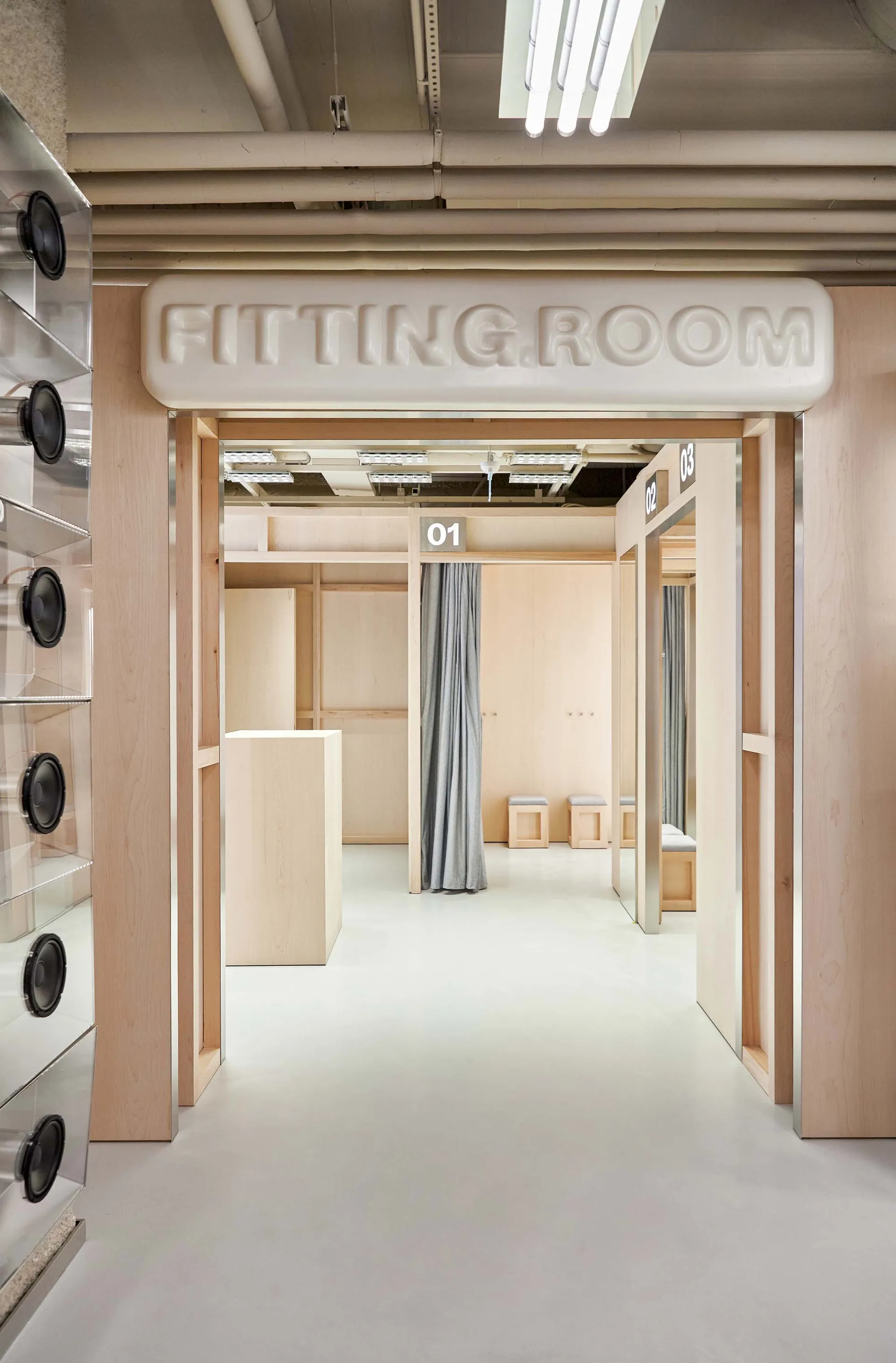 View into a modern minimalist fitting room area with light wood frames, numbered booths 01, 02, 03, grey curtains, and a white sign reading FITTING ROOM.