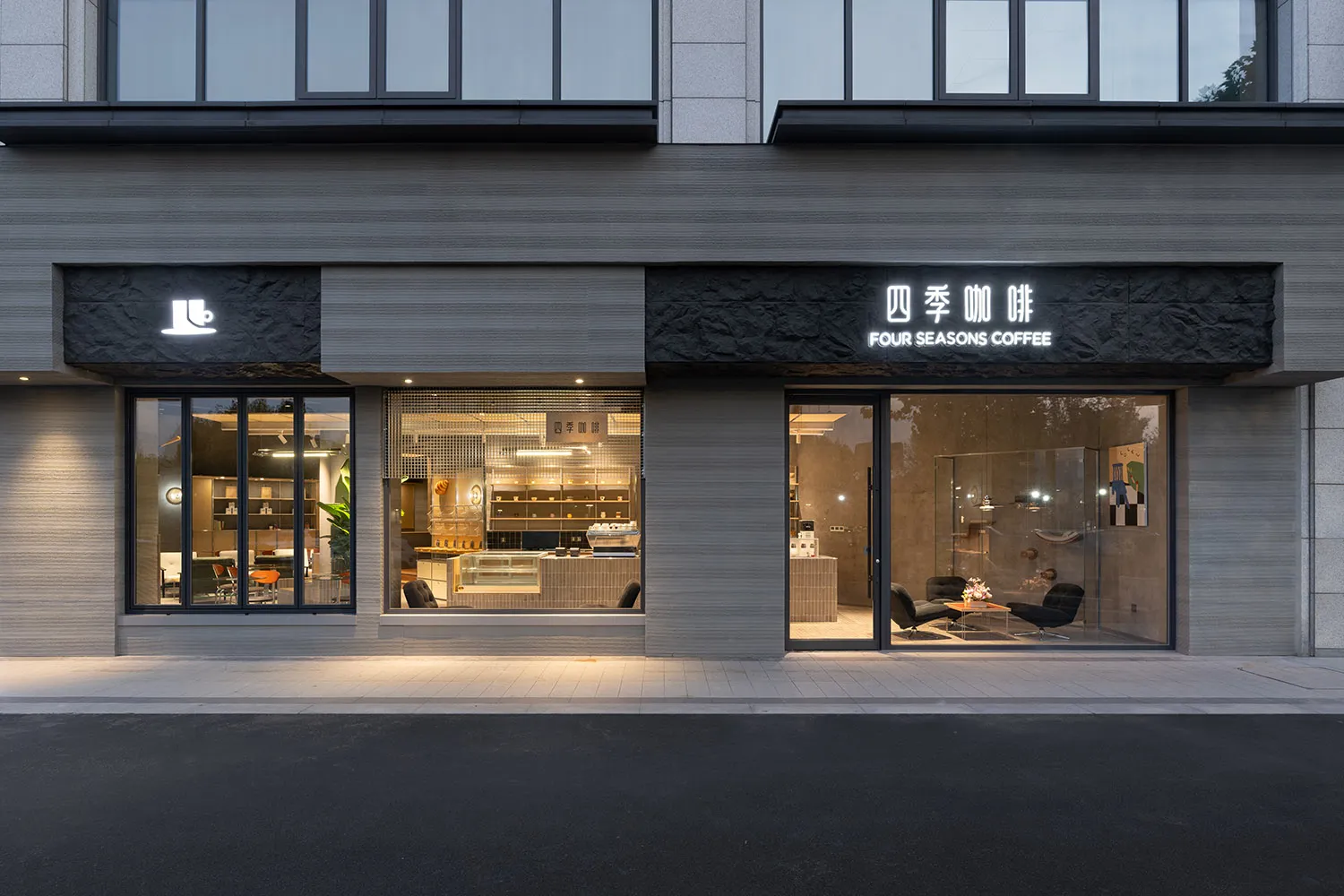 Street view of Four Seasons Coffee shop featuring a modern grey textured concrete facade, large glass windows revealing the interior, and illuminated black stone signage at dusk.