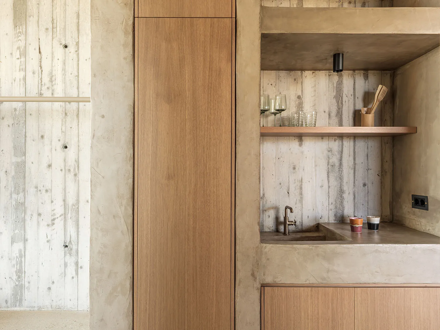 Modern minimalist kitchenette with concrete walls, oak cabinetry, integrated sink, open wooden shelf, and black faucet.