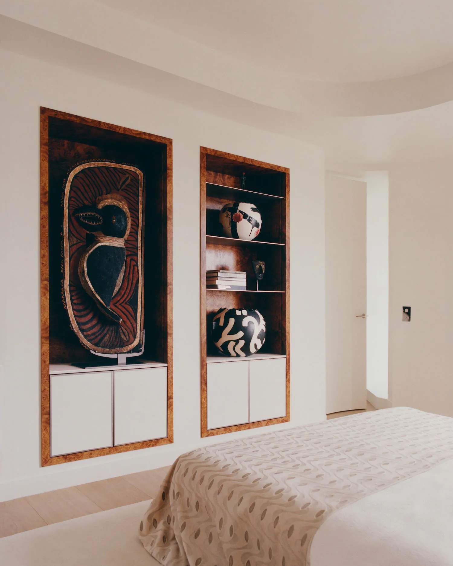 Modern bedroom featuring two recessed burl wood display niches filled with African tribal masks and pottery, white lacquered cabinetry below, and a bed with patterned beige throw in the foreground.