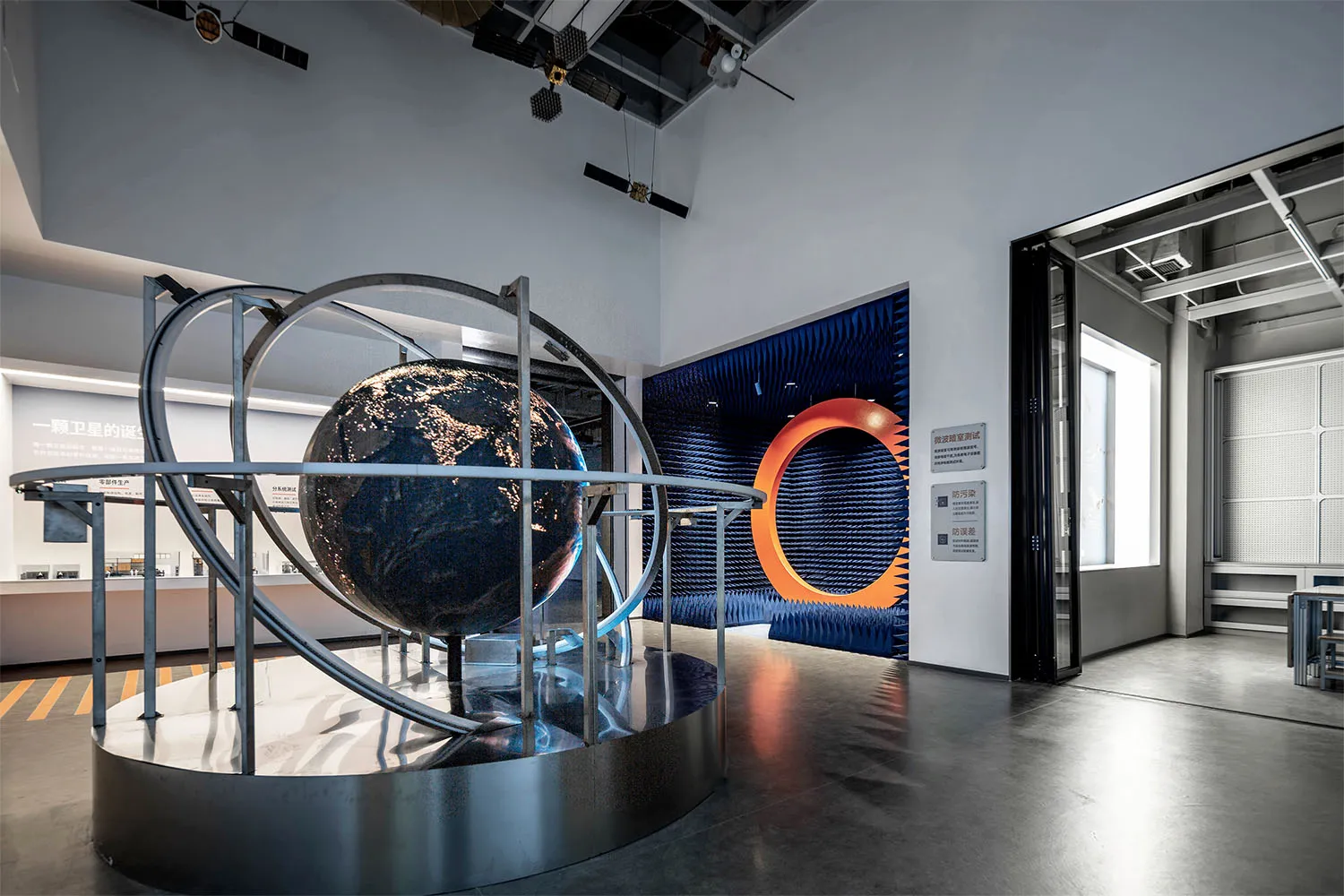 Large metallic globe exhibit with orbital rings displayed on a circular platform inside a contemporary science museum with blue acoustic panels and industrial ceiling.