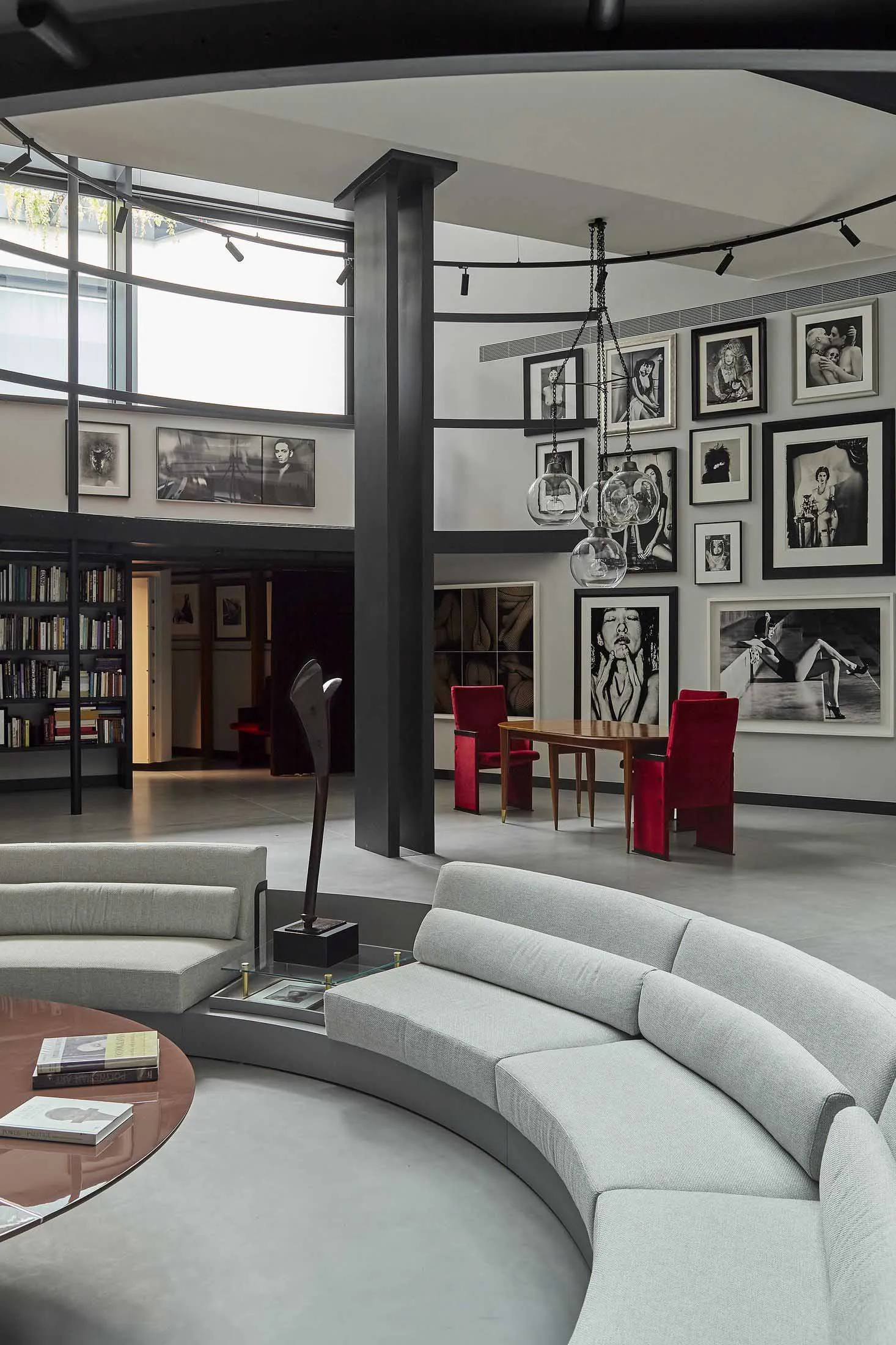 Modern double-height living room with a large curved light-grey sectional sofa, round copper coffee table, red velvet dining chairs, and walls covered in framed black and white photographs under a circular skylight.