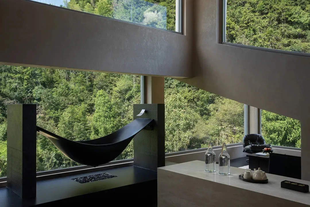 Modern concrete interior featuring a black hammock reading nook beside large windows with dense green forest view