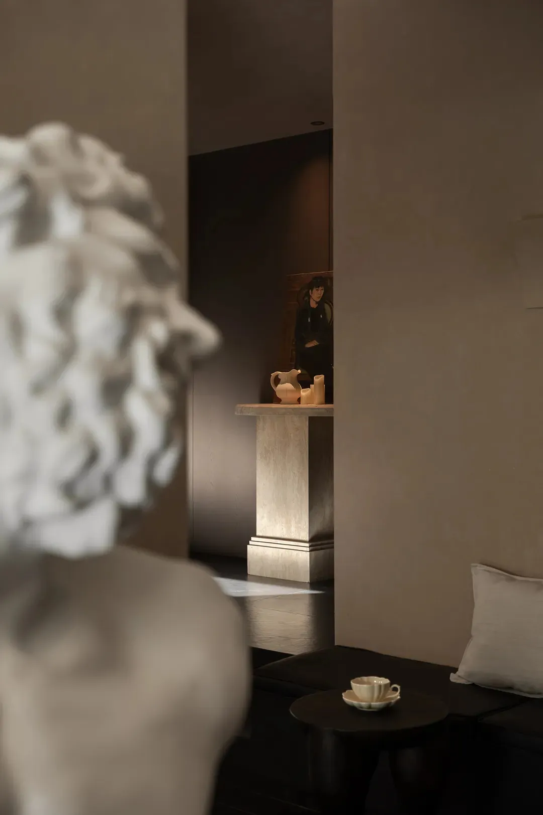 View of a contemporary minimalist living room featuring a travertine pedestal with decorative objects, a classical white marble bust in the foreground, dark flooring, and a built-in seating area with neutral cushions.