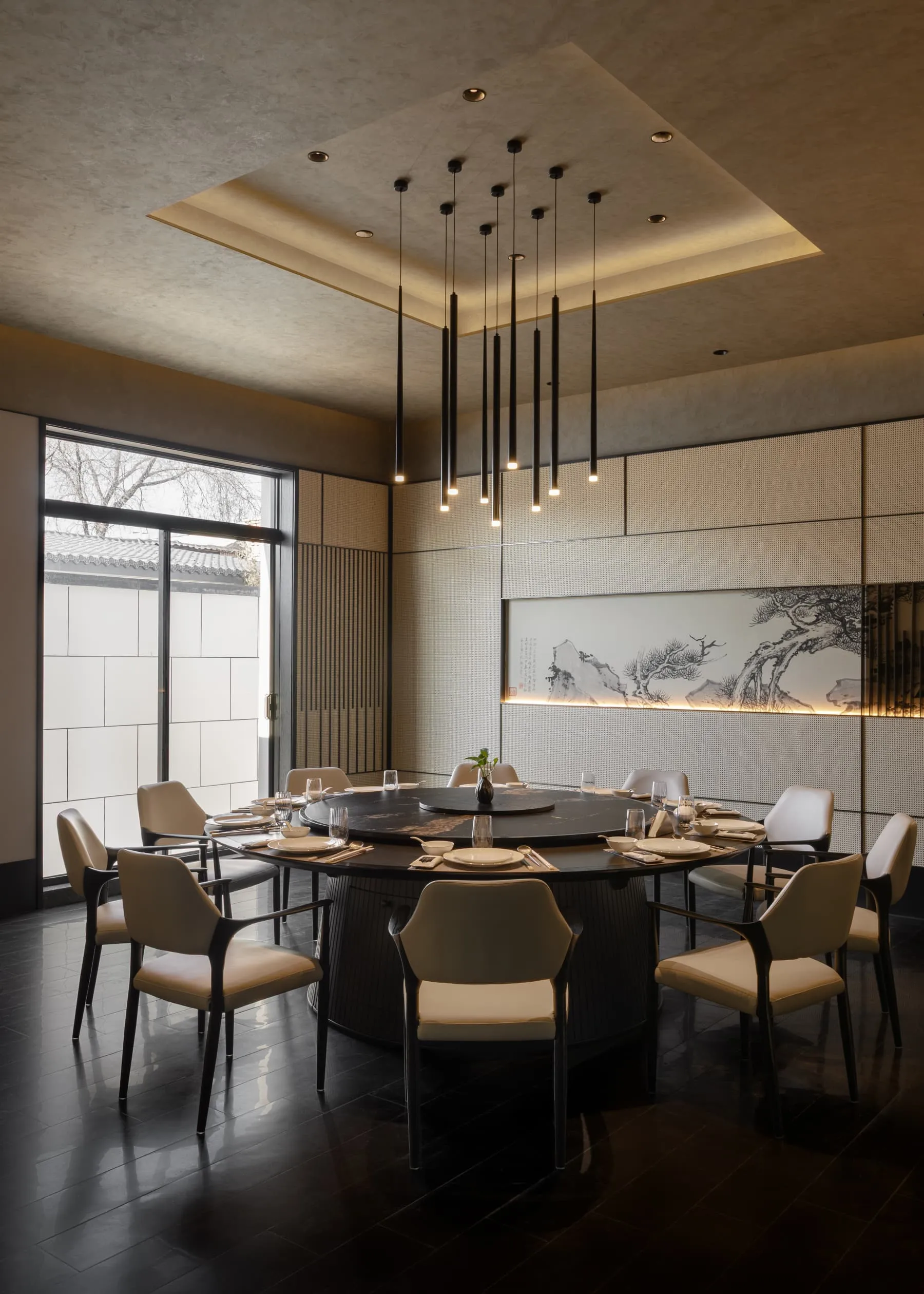 Elegant modern Chinese-style dining room featuring a large black marble round table, ten cream upholstered chairs, dramatic black pendant lights, and a backlit Chinese ink painting of pine trees and mountains.