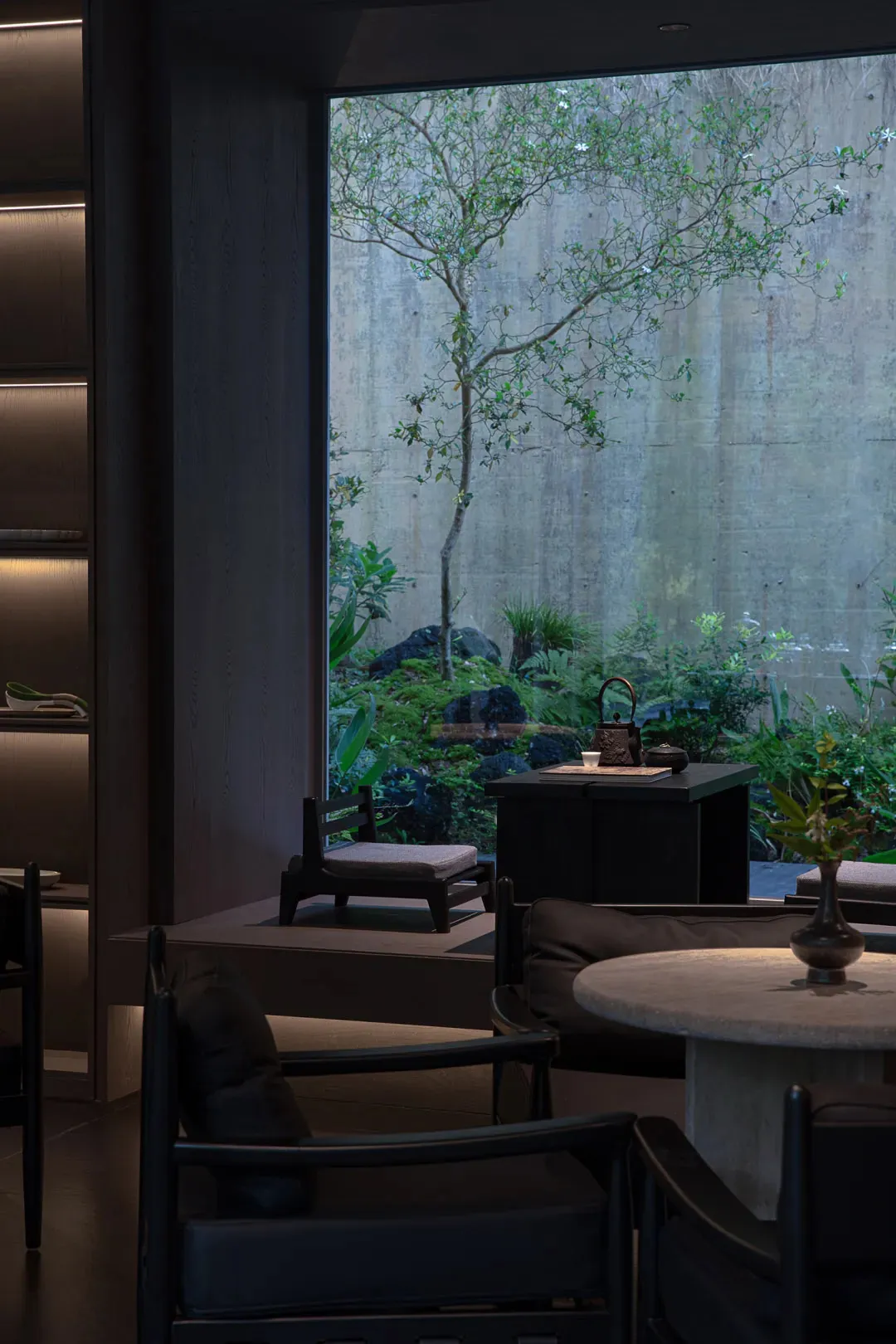 Modern Japanese-style tea room interior featuring dark wooden chairs, a round stone table, built-in seating, and a large window overlooking a lush green garden with a flowering tree against a concrete wall.