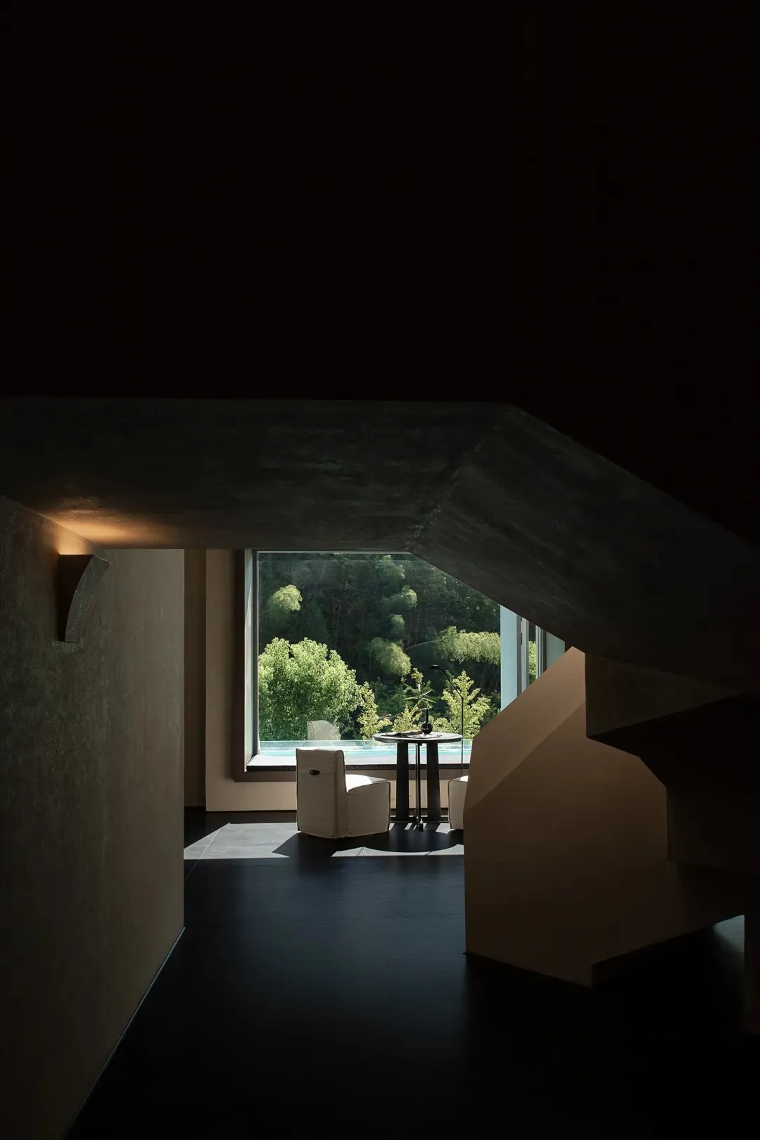 Modern minimalist living space featuring raw concrete architecture, a white armchair, round side table, and large window revealing a dense forest view.