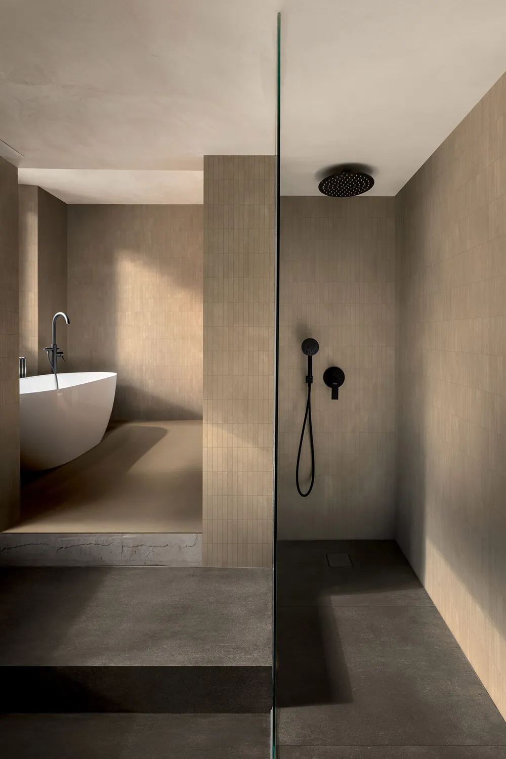 Modern minimalist bathroom featuring beige micro-cement walls and floors, a white freestanding bathtub on a raised platform, and a glass-enclosed walk-in shower with black matte fixtures.