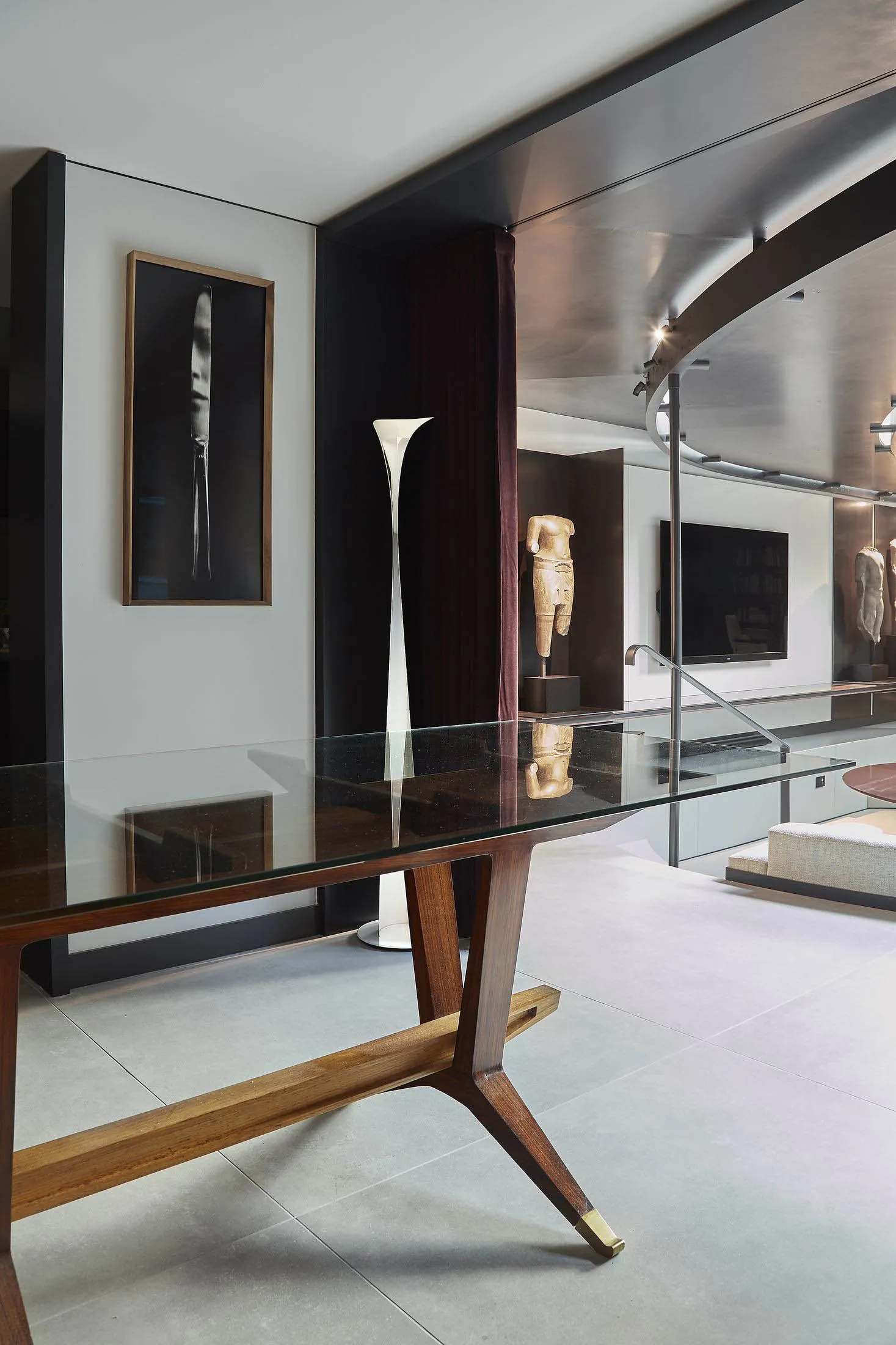 Modern living room interior featuring a walnut and glass console table, tall white floor lamp, framed black and white portrait, bronze classical torso sculptures, and a curved metallic ceiling detail.
