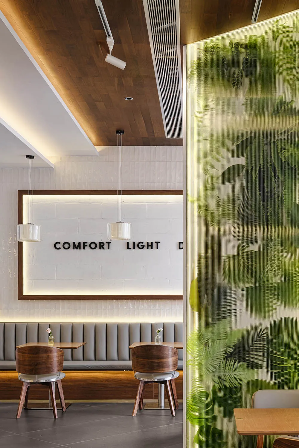 Modern cafe interior featuring walnut swivel chairs, grey upholstered booth seating, warm wood ceiling, and a glowing translucent green botanical partition wall.