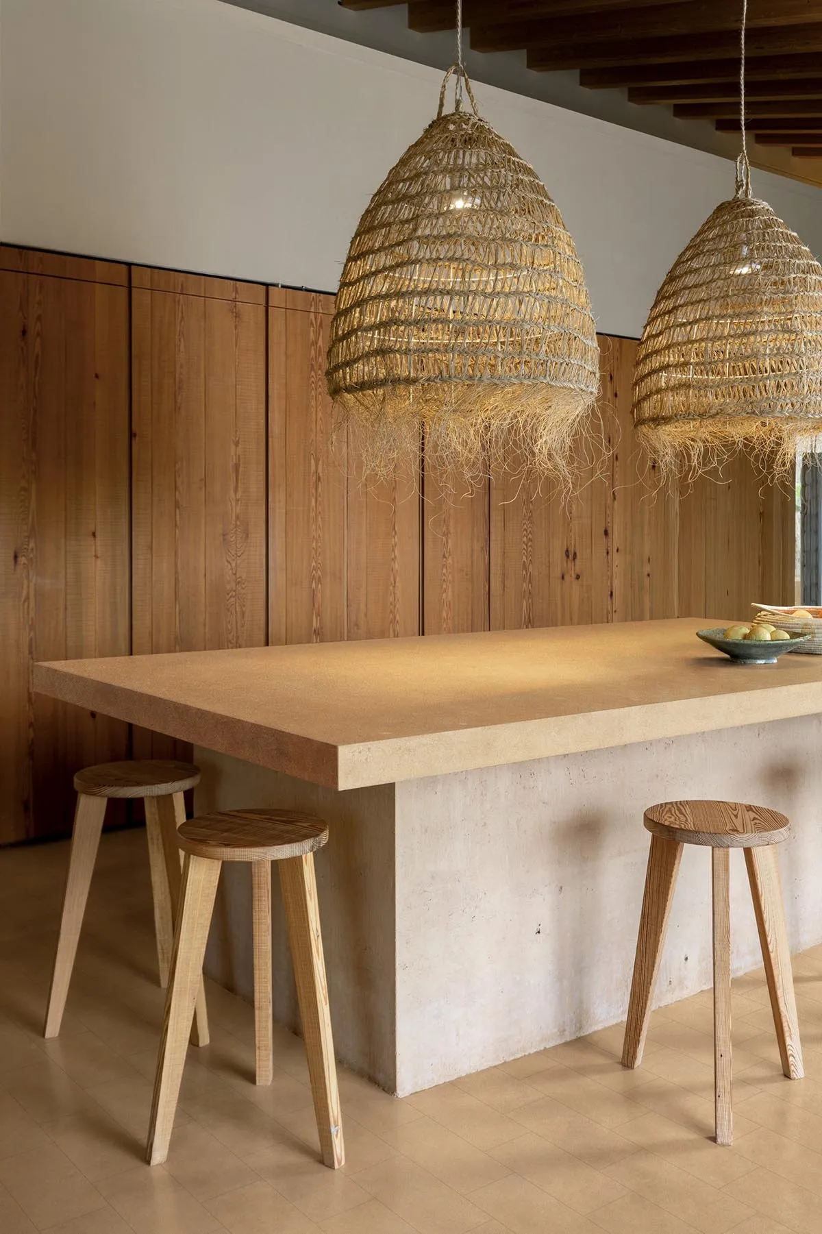 Modern minimalist kitchen island made of concrete and wood, paired with two large woven rattan pendant lights and three wooden stools against a vertical wood-paneled wall.
