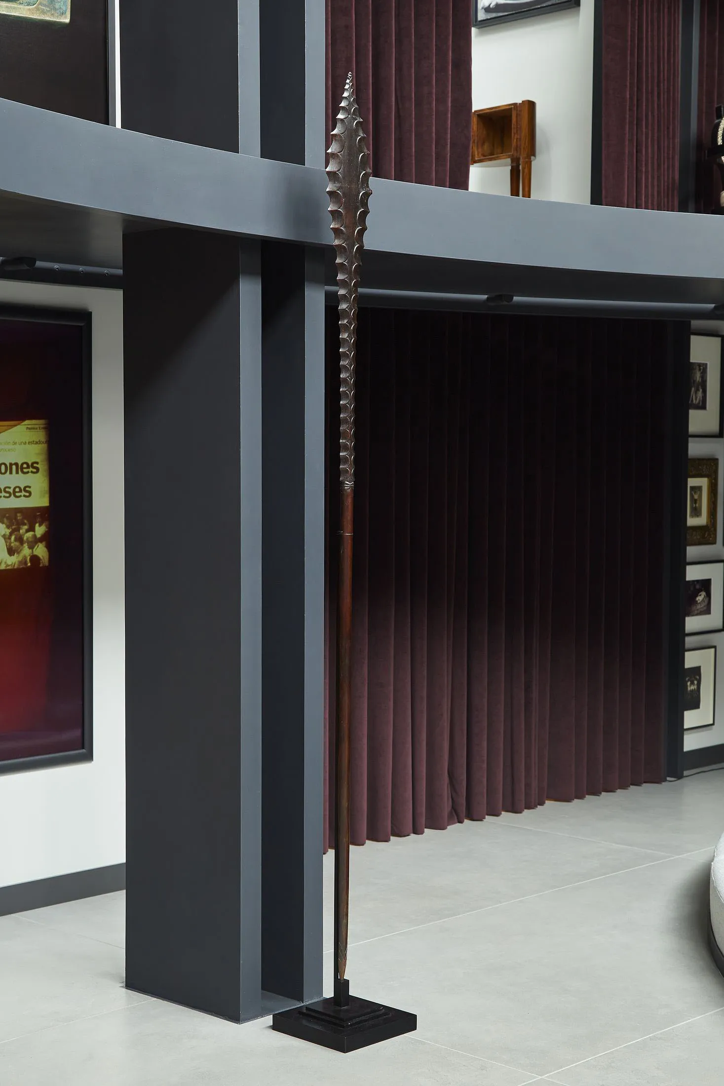 Modern living room interior featuring a tall tribal spear sculpture, dark grey structural beams, burgundy velvet curtains, and gallery-style framed art on white walls.