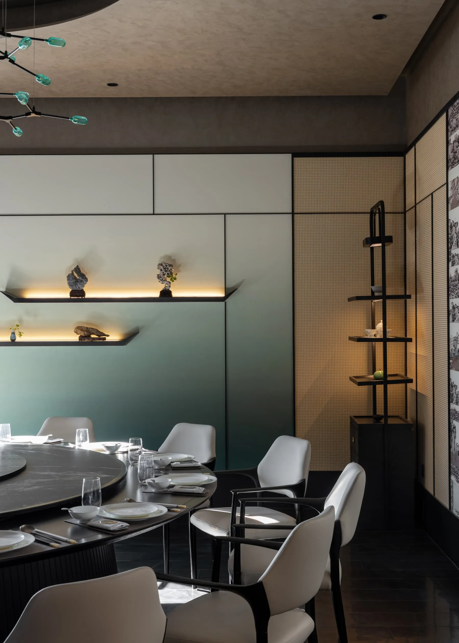 Modern dining room featuring a large round black marble table, white leather dining chairs with black frames, built-in illuminated display shelves with scholar rocks, and a teal accent wall under a modern glass chandelier.